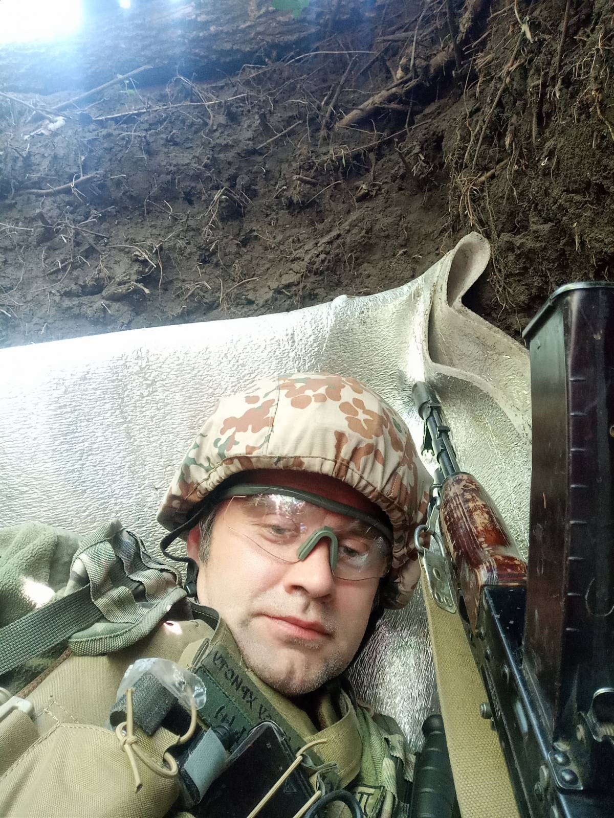 A soldier in a helmet lying in an earth dugout against a sheet of foil insulation, rifle beside him