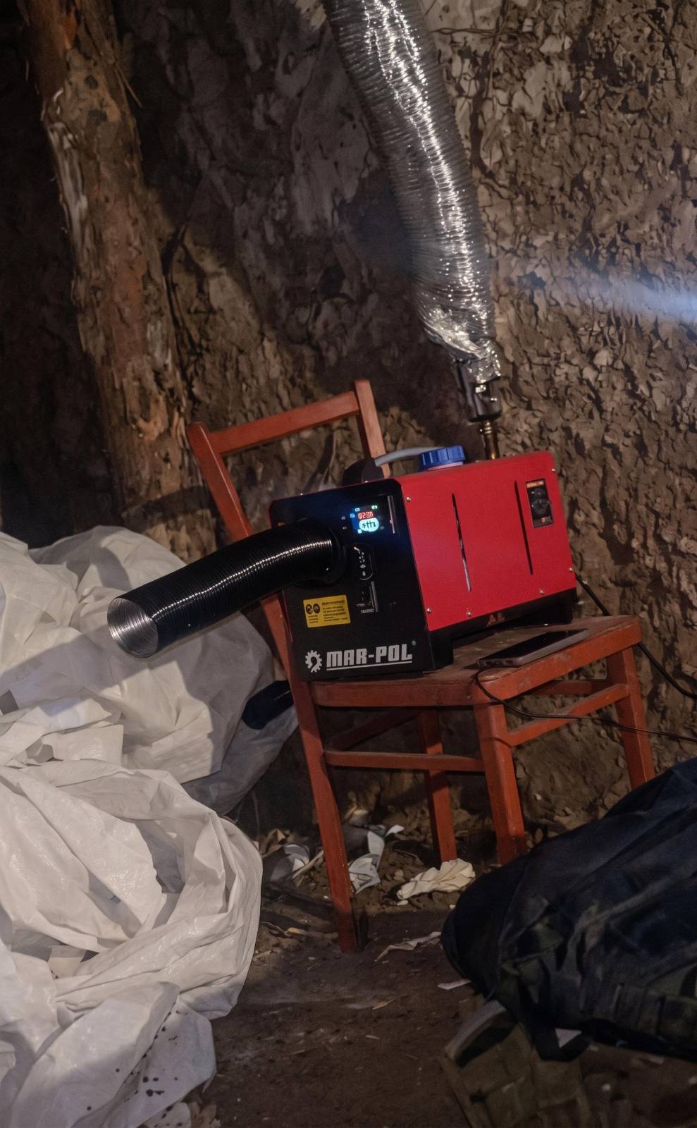 A red diesel heater running on a wooden chair inside an earth dugout, its duct pointed into the room